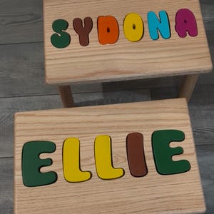 May include: Two wooden step stools with the names "SYDONA" and "ELLIE" spelled out in colorful, raised letters. The stools have a natural wood grain finish and are designed for children.