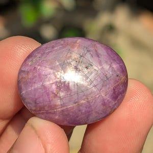 May include: A polished, oval-shaped gemstone with a deep purple hue. The stone has a reflective surface with visible internal structures and a subtle, star-like pattern. The gem is held between fingers, showcasing its size and color.