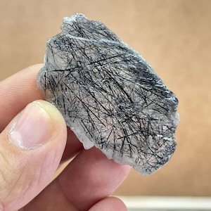 Può includere: Un cristallo grezzo trasparente con inclusioni nere a forma di ago. Il cristallo è tenuto in mano, mostrando la sua forma naturale e irregolare e il contrasto tra la base trasparente e i motivi scuri e lineari all'interno.