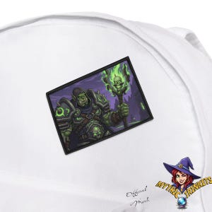 Könnte beinhalten: Weiße Mütze mit einem rechteckigen Aufnäher, der einen grünschuppigen Ork-Charakter in Rüstung zeigt, der einen Stab mit einem Totenkopf und grünen Flammen hält. Der Aufnäher ist vor einem lila Hintergrund angebracht. Die Mütze hat auch den Text "Official Merch" und ein Logo.