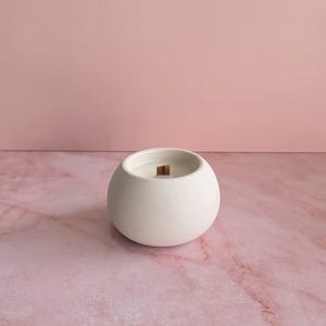 May include: A white, spherical candle in a ceramic container with a wooden wick. The candle sits on a pink marble surface against a solid pink background. The candle's design is minimalist and modern.