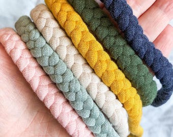 May include: A set of five braided fabric headbands in a variety of colors including soft pink, sage green, beige, mustard yellow, olive green, and navy blue. Each headband features a thick, woven texture made from soft, flexible textile material.