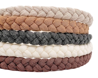 May include: A stack of five braided headbands in various neutral colors including beige, tan, charcoal gray, cream, and dark brown. Each headband features a thick, woven fabric texture with a consistent braided pattern throughout the entire band.