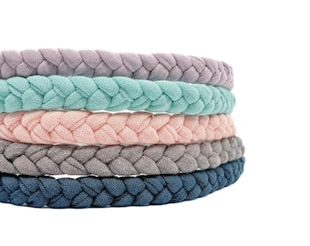 May include: A stack of five braided fabric headbands in a variety of colors, including lavender, mint green, soft pink, light gray, and dark navy blue. Each headband features a consistent three-strand plait design made from a textured textile material.