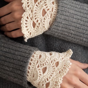 Pode incluir: Close-up de duas punhos de crochê creme com uma borda ondulada. Os punhos são usados sobre um suéter cinza. O design de crochê apresenta padrões vazados. Os punhos adicionam um toque delicado e artesanal ao traje.