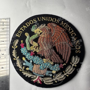 Op de afbeelding: Een ronde, zwarte geborduurde patch met het Mexicaanse wapen. Het ontwerp omvat een gouden adelaar met een slang, een cactus en de tekst "ESTADOS UNIDOS MEXICO" in goud. Een liniaal toont inches.