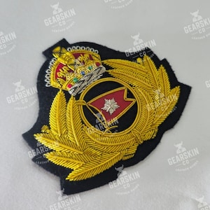 Puede incluir: Parche bordado detallado con una corona dorada, hojas de laurel y un diseño de bandera roja. El parche está sobre un fondo negro con detalles de hilo dorado. El texto "GEARSKIN CO" es visible.