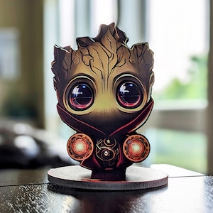 Könnte beinhalten: Eine Holzfigur eines kleinen, cartoonartigen Groot aus Guardians of the Galaxy, der einen roten Umhang trägt und die Infinity-Steine hält. Die Figur hat große, ausdrucksstarke Augen und eine detaillierte, strukturierte Oberfläche.