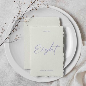 Peut inclure: Deux cartons de numéros de table blanc cassé avec des bords déchirés, avec le mot "Eight" en script violet clair. Les cartes sont sur une assiette blanche, avec une brindille de feuillage séché et le texte "Blair & Edward".