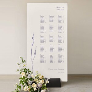 May include: An off-white wedding seating plan with names listed alphabetically. The text "PLEASE FIND YOUR SEAT" is at the top. A floral arrangement with white and yellow flowers sits in front of the chart. The names are in a dark purple font.
