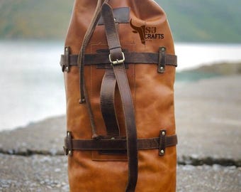 Könnte beinhalten: Eine hohe, braune Leder-Duffel-Tasche mit Kordelzugverschluss und verstellbarem Schulterriemen. Die Tasche hat dunkelbraune Lederriemen und Metallschnallen. Das Logo "SJ Crafts" ist auf der Seite der Tasche zu sehen.