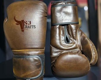 May include: A pair of brown leather boxing gloves. The gloves have a distressed finish and black accents. The logo "SJ Crafts by SQ" is printed on the left glove. The gloves are designed for boxing and other combat sports.