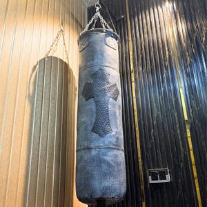 May include: A gray leather punching bag with a large black cross design. The bag is cylindrical and hangs from a chain. The bag has a textured surface and is suspended in front of a wall with vertical lines.