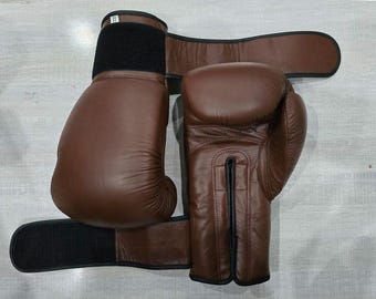 May include: A pair of brown leather boxing gloves with black wrist straps. The gloves are a deep brown colour and appear well-padded. The wrist straps have a hook and loop closure for a secure fit. The gloves are designed for boxing or other combat sports.