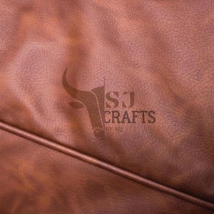 May include: Close-up of brown leather with a textured surface. The logo "SJ CRAFTS BY NQ" is visible in the center. The leather has a rich, warm tone and a subtle sheen, with a seam running across the bottom.