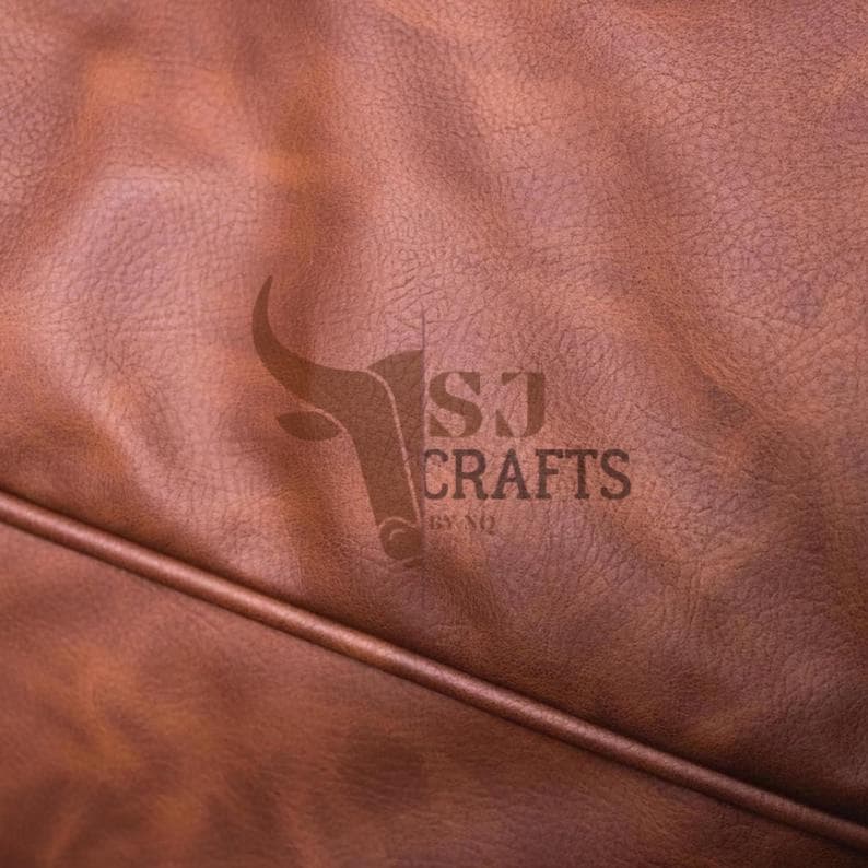 May include: Close-up of brown leather with a textured surface. The logo "SJ CRAFTS BY NQ" is visible in the center. The leather has a rich, warm tone and a subtle sheen, with a seam running across the bottom.