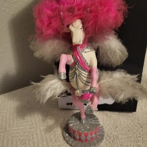 May include: A whimsical figurine of a white horse with pink accents, a pink feathery mane, and white feathery wings. The horse is adorned with silver glitter and stands on a silver and pink base. The figurine has a circus theme.
