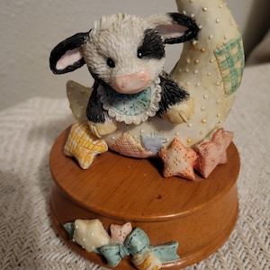 May include: A decorative figurine of a black and white cow wearing a blue bib, sitting on a crescent moon. The moon is cream-colored with polka dots and a green plaid patch. The figurine sits on a wooden base with star-shaped accents.