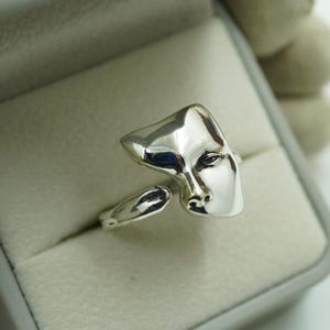 May include: A polished silver ring featuring a sculptural design that resembles a stylised human face. The metal surface is smooth and reflective, with an open band construction that allows for adjustment. The ring is presented in a box.