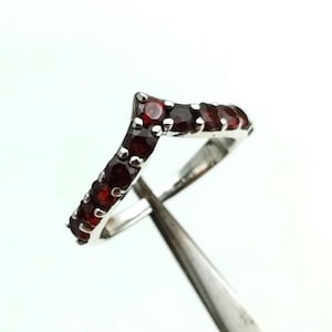 Könnte beinhalten: Ein silberfarbener Ring mit Chevron-Design, besetzt mit kleinen, runden, tiefroten Edelsteinen. Der Ring wird von einer Pinzette vor einem weißen Hintergrund gehalten. Die Edelsteine sind gleichmäßig auf der gebogenen Oberfläche des Rings angeordnet.