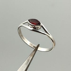 May include: A silver ring featuring a deep red oval gemstone. The ring has a delicate, minimalist design with a split band that curves up to cradle the stone. The gemstone is set in a simple bezel.
