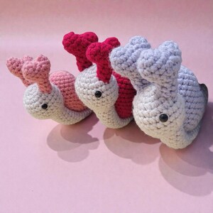 May include: Three handmade crochet snails in a row. The snails have white bodies, with pink, red, and gray shells. Each snail has black button eyes and a heart-shaped head. The background is a soft pink.