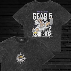 May include: A charcoal gray acid-wash t-shirt featuring a small compass and skull graphic on the front. The back displays a large graphic of Monkey D. Luffy in Gear 5 form with the text "GEAR 5" and the "ONE PIECE" logo in white and purple.