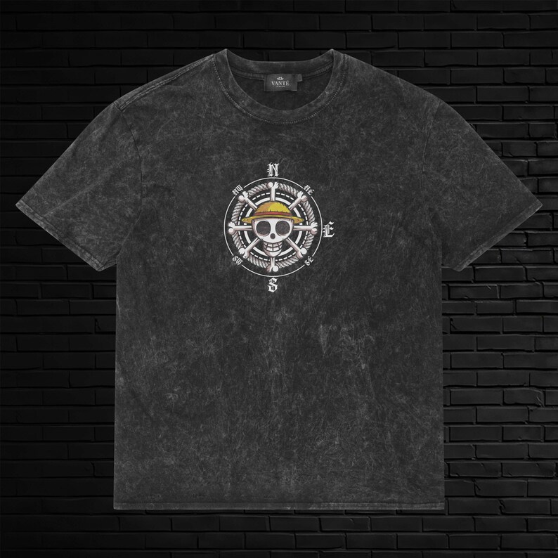 May include: A charcoal gray acid-wash t-shirt featuring a central graphic of a skull wearing a yellow straw hat, positioned over a ship's wheel with compass directions N, E, S, and W. The shirt has short sleeves and a crew neckline.