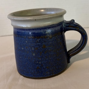 May include: A ceramic mug featuring a deep blue body with a textured, speckled finish and a smooth, light gray rim. The mug has a sturdy, curved handle attached to the side, with a distinct flared lip at the top edge and a dark base.