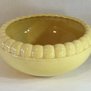 May include: A small, round, yellow ceramic bowl with a scalloped rim. The bowl has a smooth, glossy finish and a simple design. The interior of the bowl is a slightly darker shade of yellow. The bowl is likely 10-15 cm in diameter.