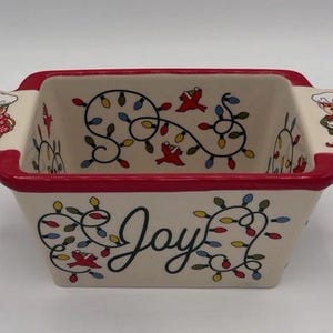 May include: A rectangular ceramic baking dish with a red rim and handles. The dish is decorated with a Christmas theme, featuring string lights, red birds, and the word "Joy". Gingerbread figures are on the handles.