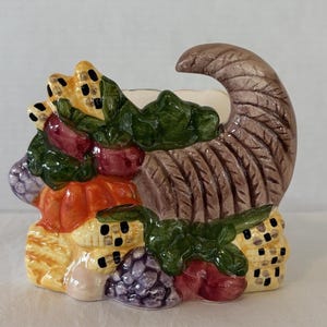 May include: A ceramic cornucopia overflowing with colorful, sculpted fruits and vegetables. The horn is brown with textured details, and the arrangement includes orange pumpkins, purple grapes, and green leaves. A decorative piece for the fall season.