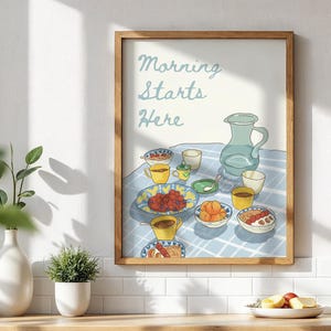 Puede incluir: Un cuadro enmarcado con el texto "Morning Starts Here" sobre una escena de desayuno. La ilustración presenta una jarra azul, tazas y platos de comida sobre un mantel a cuadros. El marco es de color marrón claro.