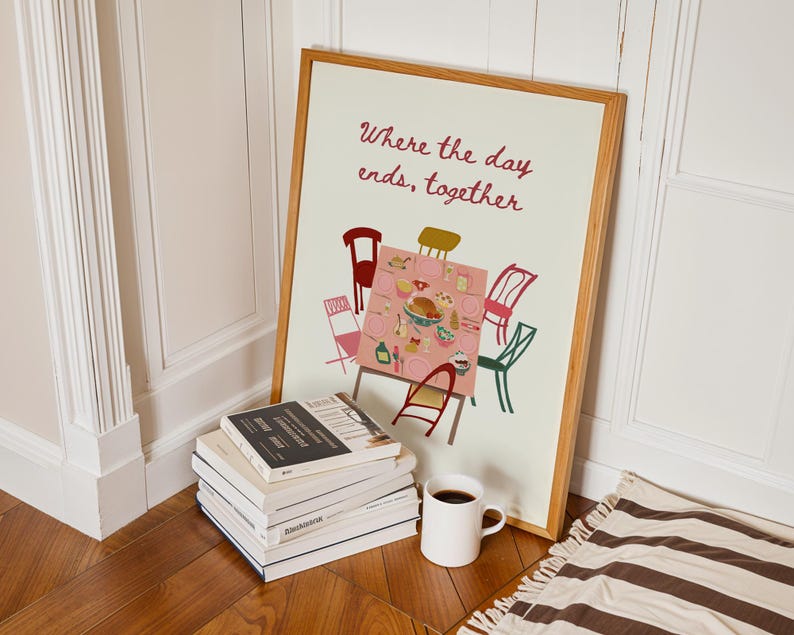 Puede incluir: Una impresi&oacute;n art&iacute;stica enmarcada con el texto "Where the day ends, together". La impresi&oacute;n representa una mesa con sillas. Una pila de libros y una taza blanca con caf&eacute; est&aacute;n frente a la impresi&oacute;n. Una alfombra a rayas est&aacute; en el suelo.