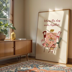 Puede incluir: Una impresi&oacute;n art&iacute;stica enmarcada que presenta una escena de mesa de comedor con sillas de varios colores. El texto "Where the day ends, together" se muestra en cursiva. La obra de arte est&aacute; apoyada contra una pared cerca de un mueble de madera y una ventana.