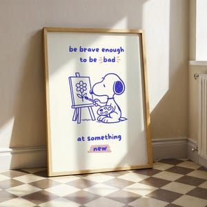 May include: Framed art print featuring a blue line drawing of a cartoon dog painting a flower on an easel. The text reads "be brave enough to be bad" and "at something new". The artwork is in a wooden frame.