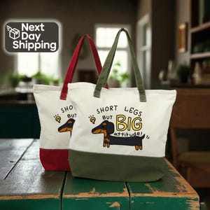 May include: Two canvas tote bags with white upper sections and colored bottoms. One has a red bottom and handle, the other has a green bottom and handle. Each bag features a dachshund graphic and the text "Short Legs But Big Attitude!"