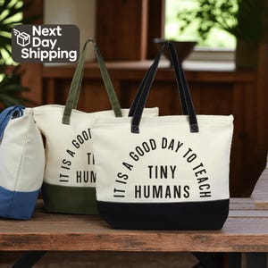 May include: Canvas tote bags with black and olive green accents. The bags have black handles and the text "IT IS A GOOD DAY TO TEACH TINY HUMANS" printed on the front. The bags are on a wooden surface.