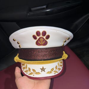 Può includere: Un berretto da capitano bianco con un emblema a forma di zampa color marrone scuro contenente un'ancora dorata. Il berretto presenta una fascia glitterata marrone scuro, un cordoncino intrecciato dorato e una visiera bianca con foglie d'oro.