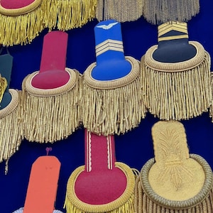 Puede incluir: Una colección de charreteras militares en varios colores, incluyendo rojo, azul, negro y dorado. Cada charretera presenta intrincadas borlas doradas y elementos decorativos, exhibidas sobre un fondo azul oscuro. Las charreteras varían en forma y diseño.