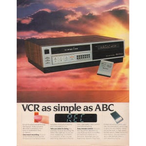 May include: A vintage VCR with a wood-grain finish, silver accents, and a digital display. The remote control sits beside the unit. The text "VCR as simple as ABC" is displayed below the VCR.