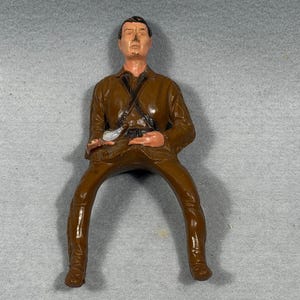 May include: A molded plastic figurine of a seated person wearing a brown jacket with a dark brown cross-body strap and matching brown trousers. The figure has a painted face with dark hair, a light skin tone, and a small white object held in the lap.