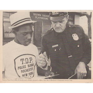 May include: Black and white photograph of a police officer and a young man. The young man wears a white t-shirt with "T.O.P. POLICE BUREAU ROCHESTER NEW YORK" printed on it. The officer is in uniform. The background shows "BROWN SQUARE PLAYGROUND".