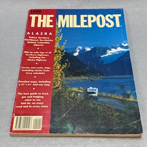May include: The 1990 edition of The Milepost travel guide features a red vertical sidebar with white text listing Alaska, Yukon Territory, Northwest Territories, British Columbia, and Alberta. The cover shows a white camper on a road near a lake.