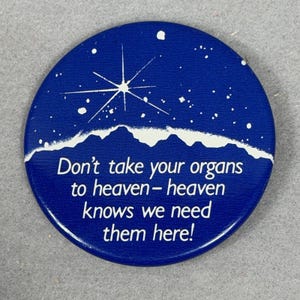 May include: A circular blue pinback button featuring a white graphic of a mountain range under a starry night sky with a large starburst. The text reads: Don't take your organs to heaven - heaven knows we need them here! The design is printed in white.