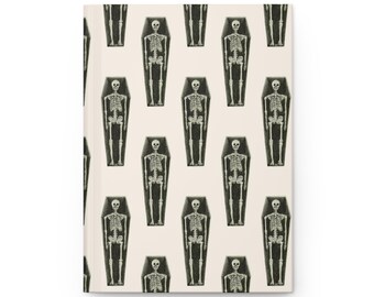 May include: A hardcover journal with a repeating pattern of skeletons in coffins. The skeletons are white, and the coffins are black. The background is a light beige color. The journal is closed and has a clean, minimalist design.