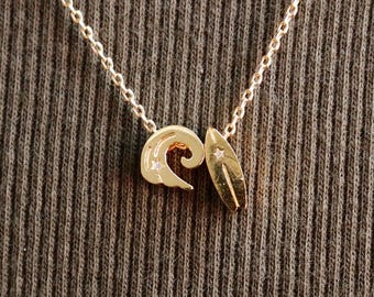 May include: A gold-colored necklace with a delicate chain. The pendant features a wave and a surfboard design. The necklace is displayed on a ribbed, dark gray fabric background. The text "emma's gems" is in the upper center.