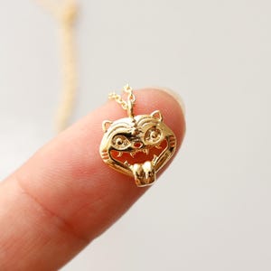 Peut inclure: Un collier avec un pendentif doré représentant une tête de tigre stylisée. Le pendentif est petit, avec des détails complexes, et est suspendu à une fine chaîne dorée. Le texte "emma's gems" est visible.