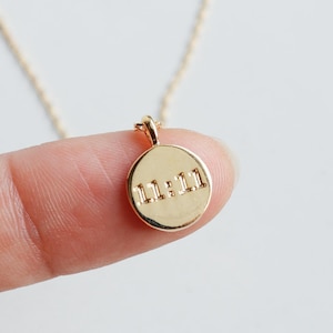 May include: A gold-colored pendant necklace with a circular charm. The charm features the engraved numbers "11:11". The necklace is held by a finger against a white background. The text "emma's gems" is in the upper right corner.