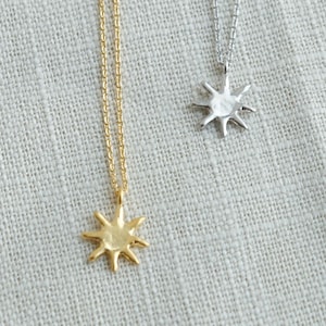 May include: Two delicate necklaces with sunburst pendants. One necklace is gold-toned, and the other is silver-toned. The pendants have a textured, spiky design. The necklaces are displayed on a light gray, textured fabric.